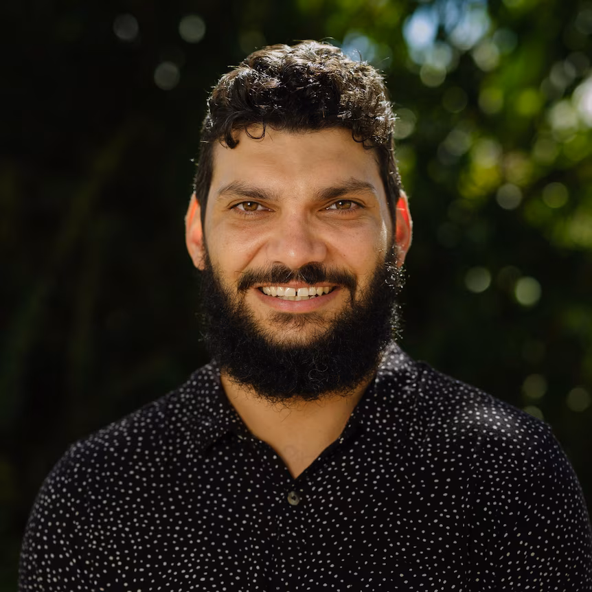 Clinton Benjamin is smiling at the camera wearing a black shirt with white spots. He has dark brown hair and a beard. He is quoted in this post about a High Court native title decision.