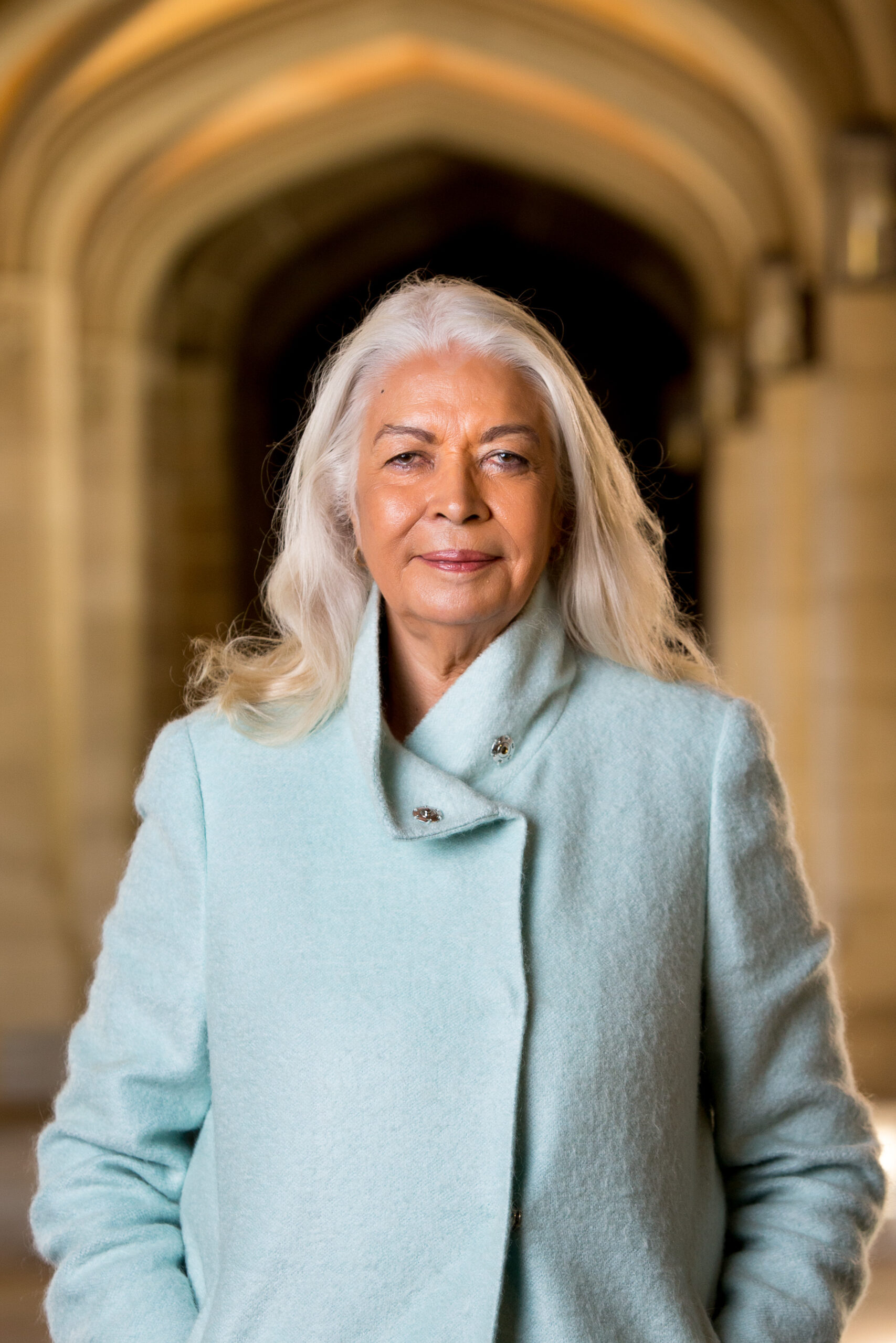 Portrait of an older woman with silver hair wearing a light blue coat in a stone archway background.