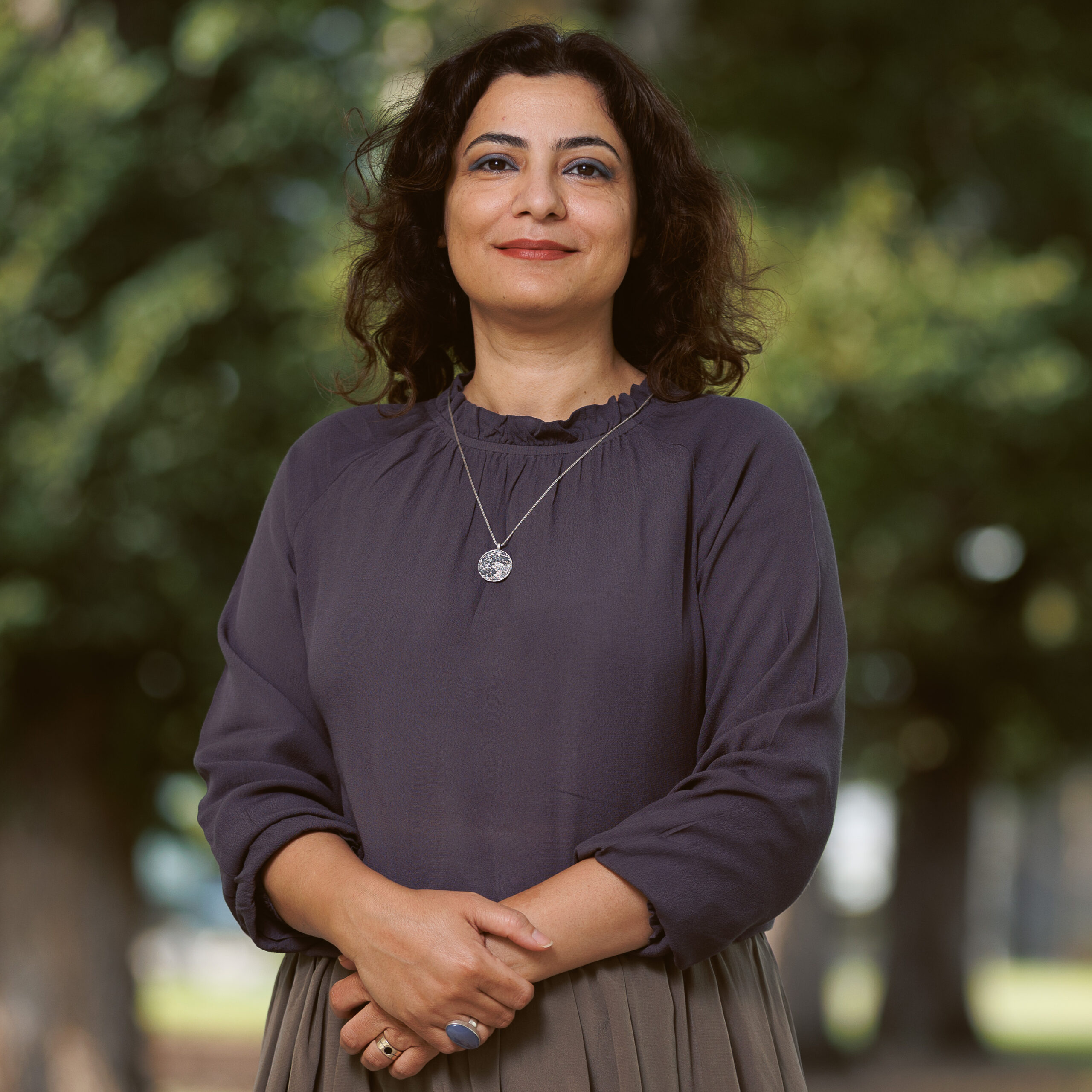 Portrait of a smiling woman outdoors in a dark purple blouse with arms crossed, wearing rings and a pendant necklace in a park setting