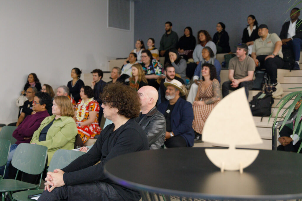 Audience seated on tiered bleachers in a bright room, attentively watching a presentation or speaker off-camera, diverse ages and styles visible.
