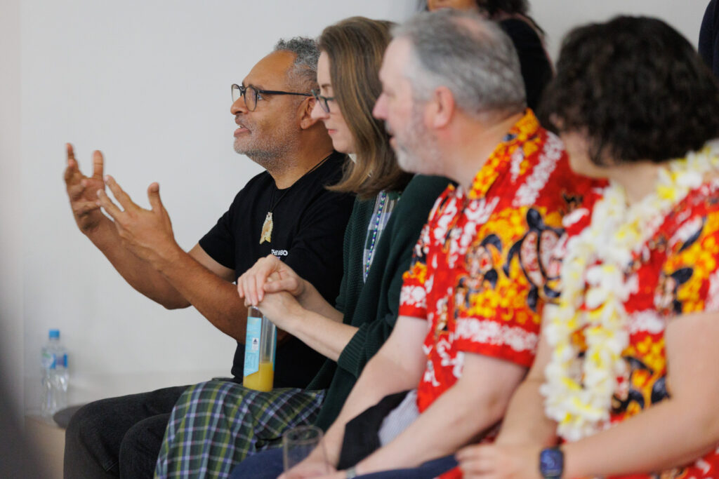 Group of people seated in a meeting; man with glasses gestures with his hands while speaking.