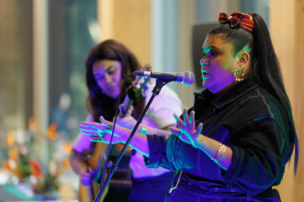 Female vocalist at a microphone on stage, with colorful stage lighting and other musicians blurred in the background.