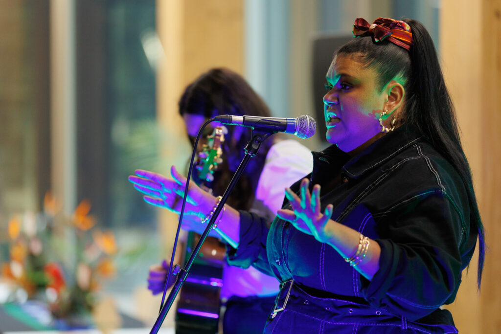 Female singer at a microphone on stage, hands raised, wearing a dark denim jacket and a colorful headband with purple lighting.