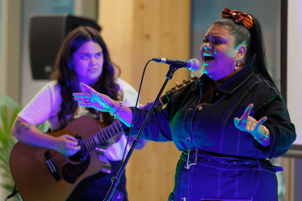 Two musicians performing onstage: a female singer at the microphone in front, with a guitarist playing in the background under colored lighting.