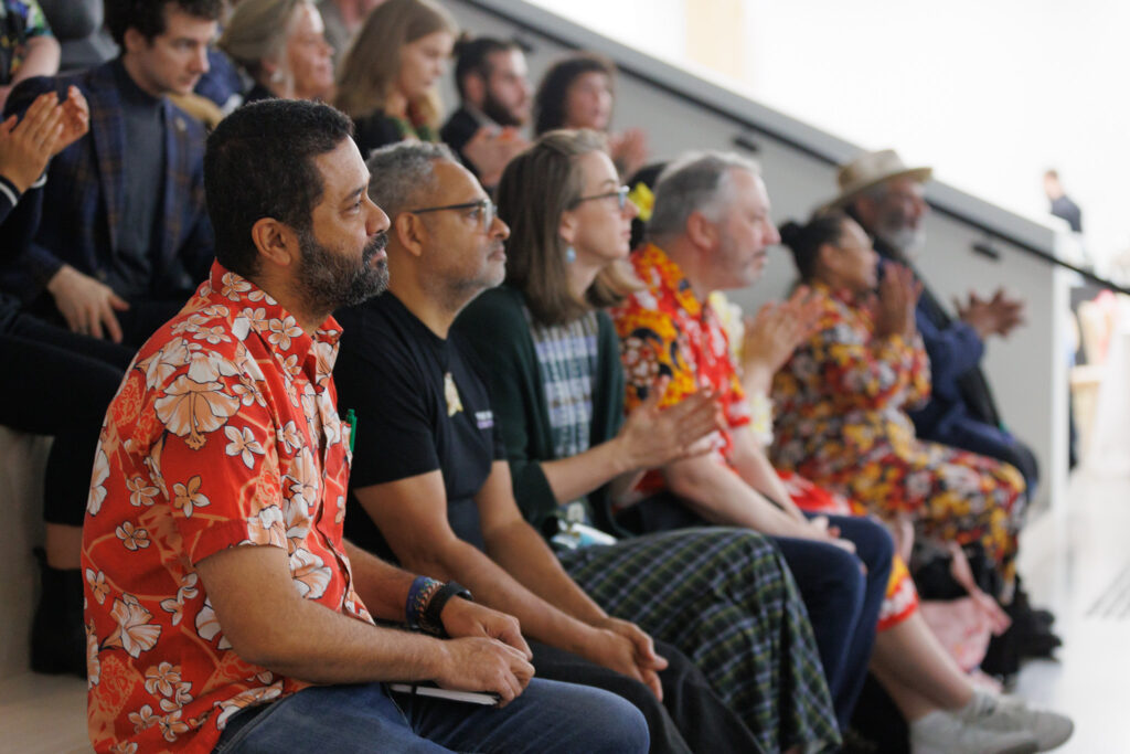 Audience members seated in a row, some wearing Hawaiian shirts and leis, watching a performance or presentation attentively.