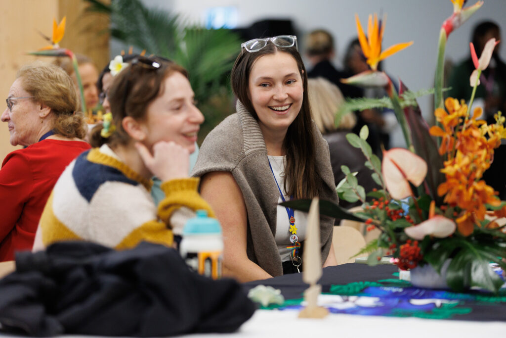 Smiling woman with glasses on her head wears a beige wrap at a floral event, with bright tropical flowers nearby.