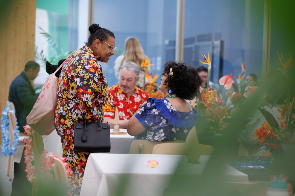 Two women in bright tropical-print dresses chat at a table during a festive gathering with flower decor in the foreground and background.