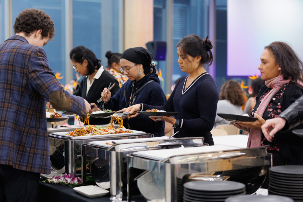 People serve themselves at a buffet line with trays of noodles and vegetables in front of them.