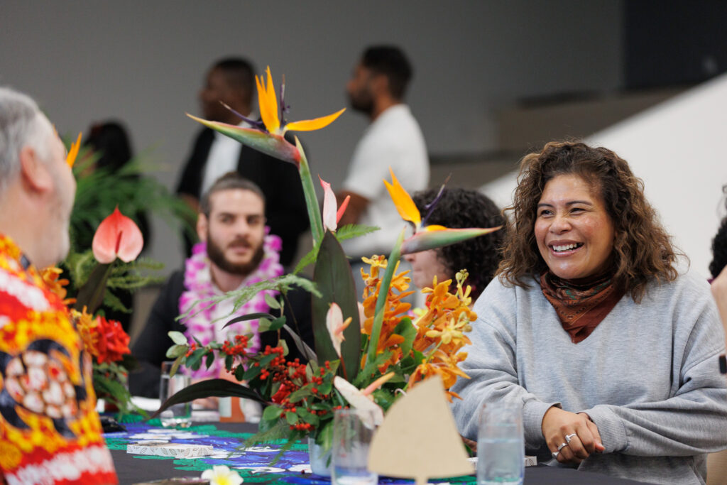Smiling woman in a gray sweatshirt sits at a table decorated with bright tropical flowers.