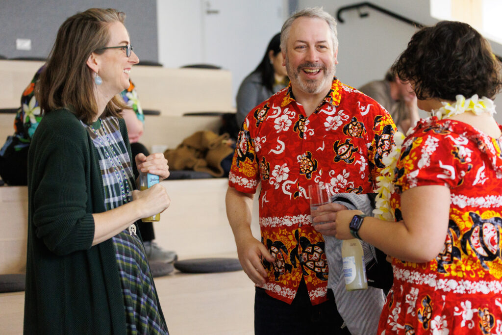 Three adults chat and laugh at a casual indoor gathering; a man in a red Hawaiian shirt holds a drink in front of him, smiling broadly.