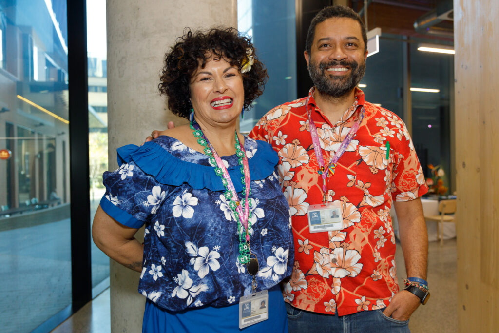 Two smiling coworkers in tropical shirts with badges posing indoors near a concrete pillar.