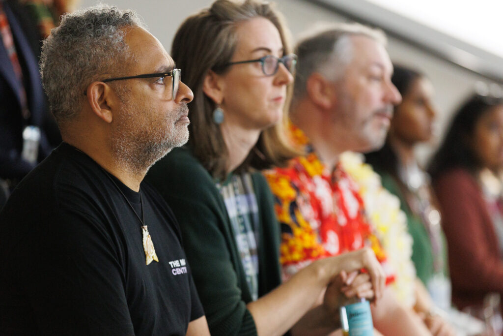 Close-up of a man with gray stubble and glasses in a black shirt, watching attentively with others wearing leis at an event.