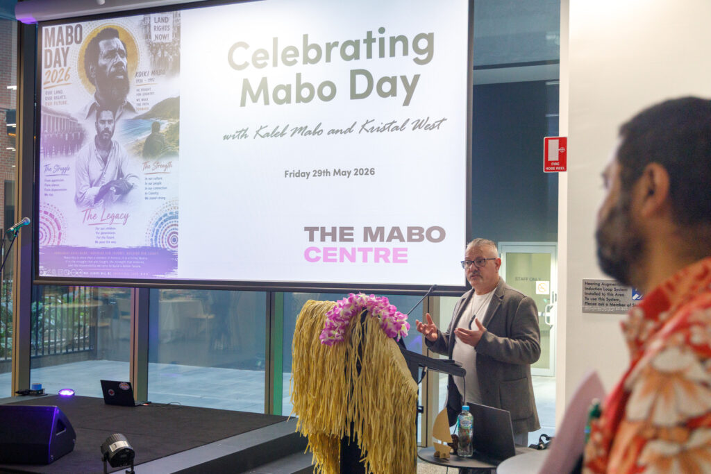Speaker at a podium on a stage with a large screen reading 'Celebrating Mabo Day' and a left-side poster about Mabo Day 2026; audience member in a colorful woven costume in the foreground.