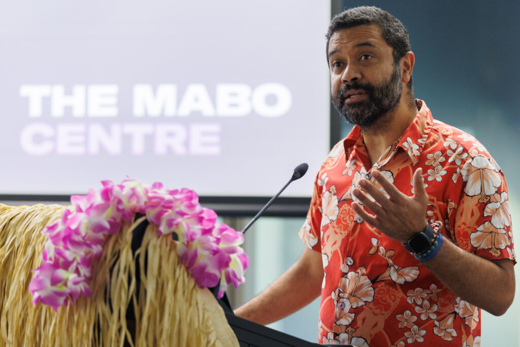 Bearded man in a red floral shirt speaks at a podium with a microphone, pink flowers in the foreground and a blurred screen in the background.