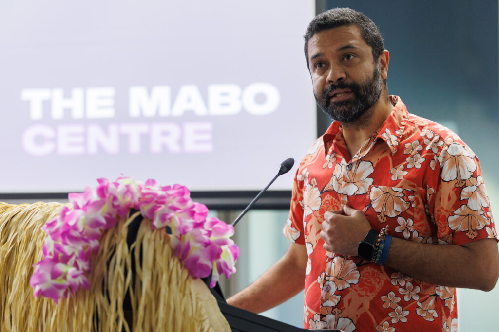 Man in a red floral Hawaiian shirt speaks at a podium with a microphone; pink flowers in the foreground and a blurred sign reading 'THE MABO CENTRE' in the background.