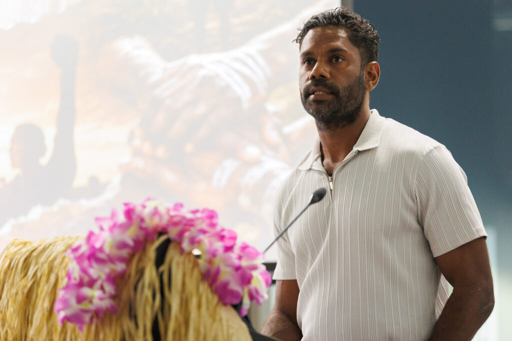 Man in a light striped polo speaking at a podium with a microphone, blurred pink flowers in the foreground.