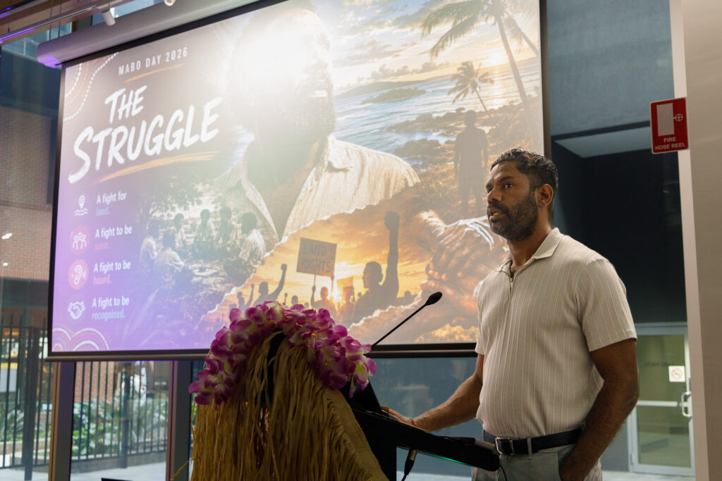 Male speaker at a podium presenting, with a large projection behind showing 'The Struggle' and tropical imagery.