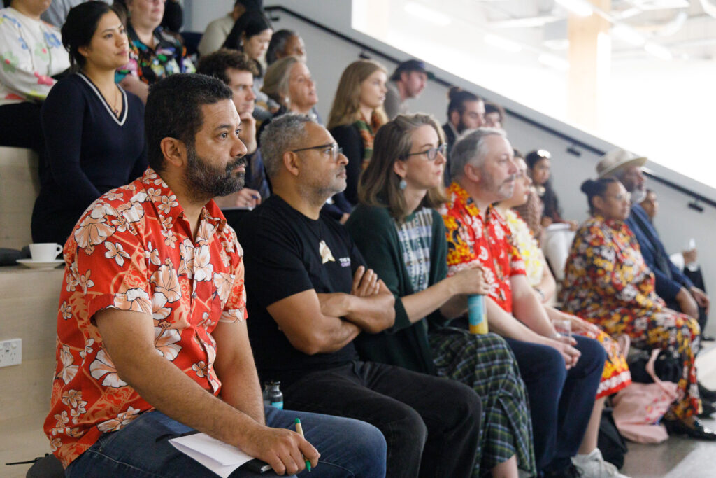 Audience of diverse adults seated on bleachers at an indoor event, many in bright tropical shirts, watching intently.