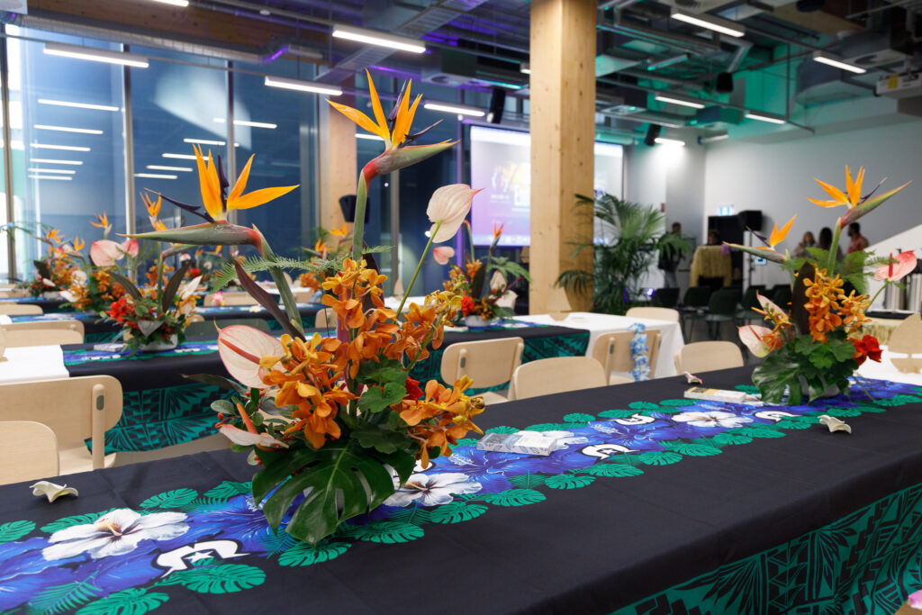 Conference room set for an event with vibrant tropical flower centerpieces on long tables and attendees in the background.