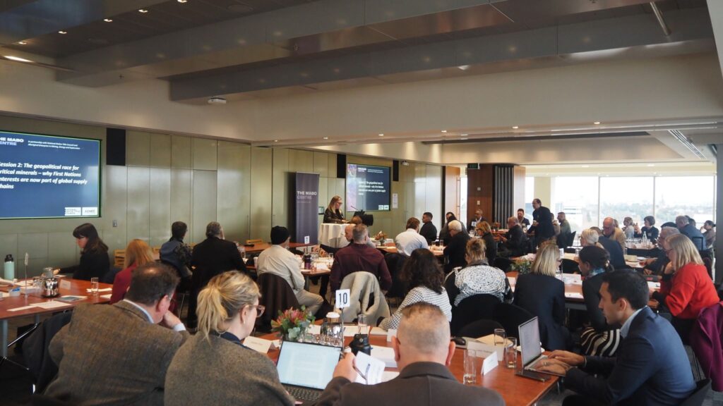 1782737006649 – The Mabo Centre Conference room with a speaker at a lectern presenting to a large group around round tables and screens displaying slides at the front.